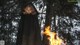 A woman in a black cloak standing in front of a fire.